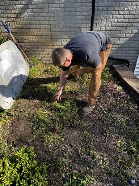 Person bending over weeding a patchy backyard garden beside a brick wall and driveway.