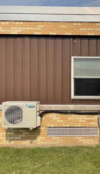 Air conditioner unit mounted on a brick and brown siding exterior wall beneath a window.