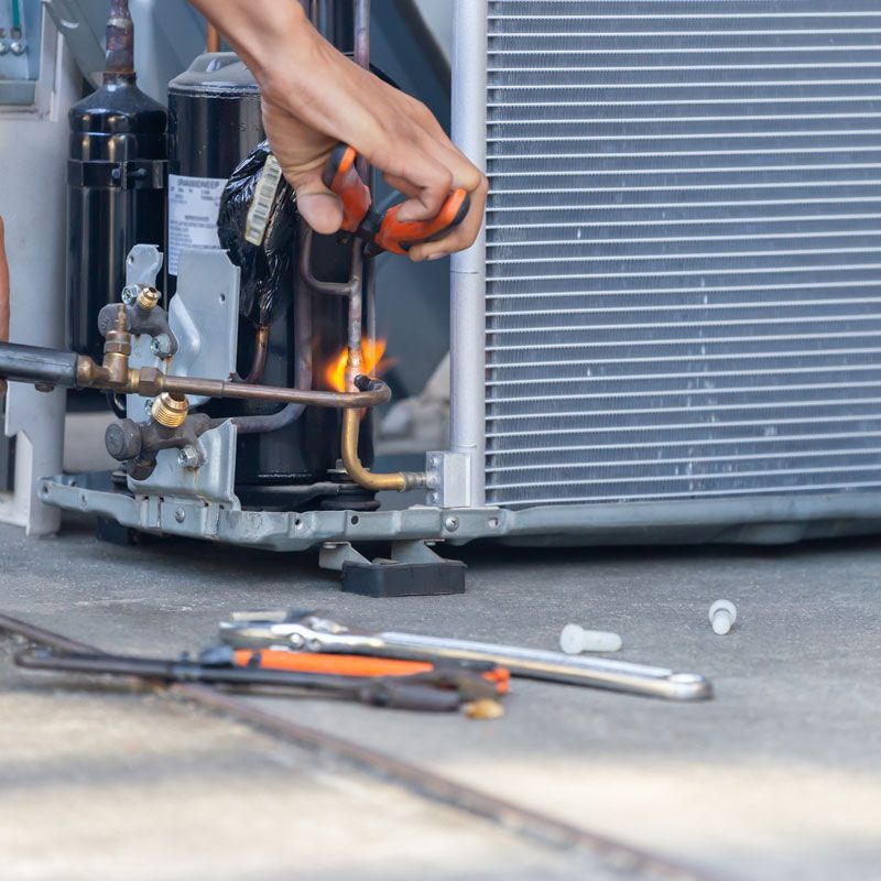 Hand repairing an HVAC unit with a torch beside tools on a concrete floor