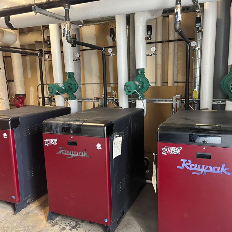 Three red and black boiler units in a basement mechanical room with pipes and valves overhead