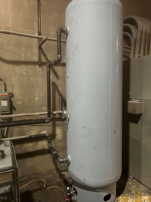 Tall white cylindrical tank in an industrial utility room with pipes and equipment nearby