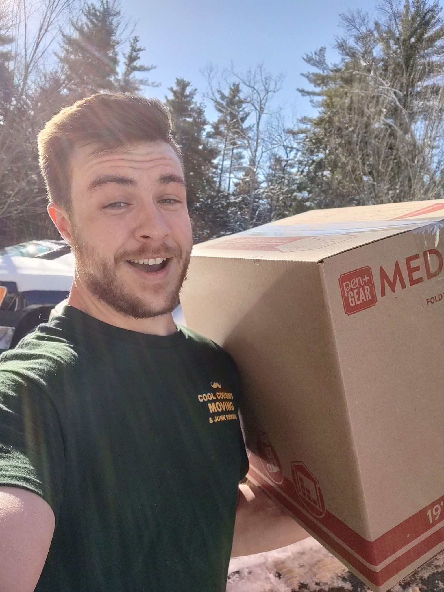 Man outdoors in sunlight carrying a large cardboard box with red markings and smiling at the camera.