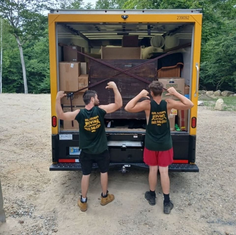 Two people flexing in front of an open moving truck packed with furniture and boxes.