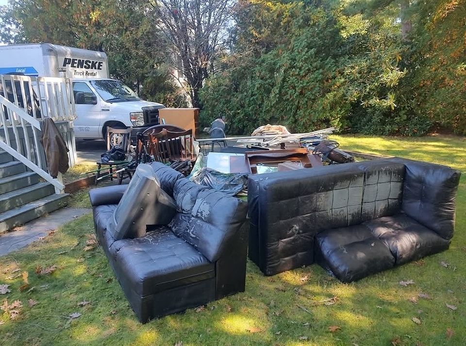Black faux-leather sectional and cushions on a lawn beside moving truck and house stairs.