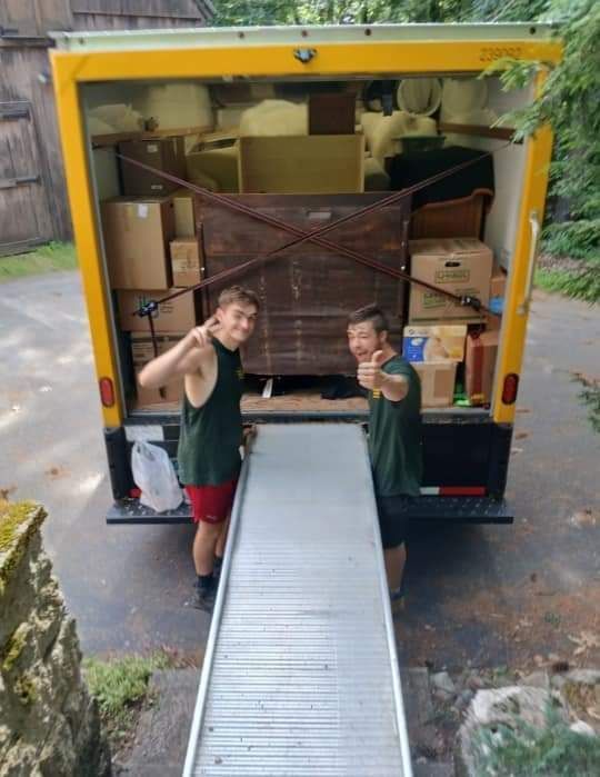 Two people unloading boxes from a yellow moving truck using a metal ramp, both smiling and giving thumbs-up.