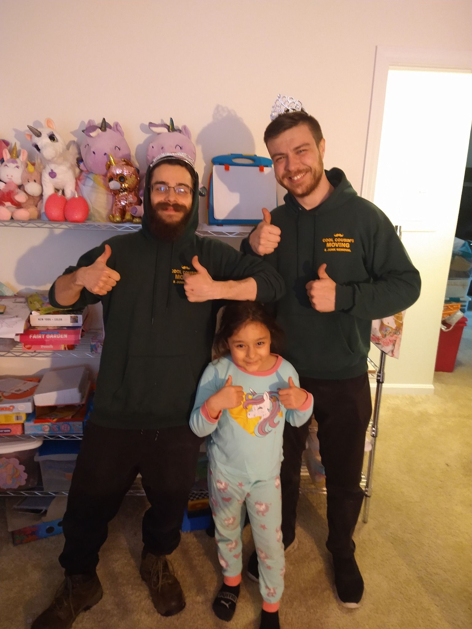 Three people smiling indoors, posing with thumbs up in a bedroom-like room.