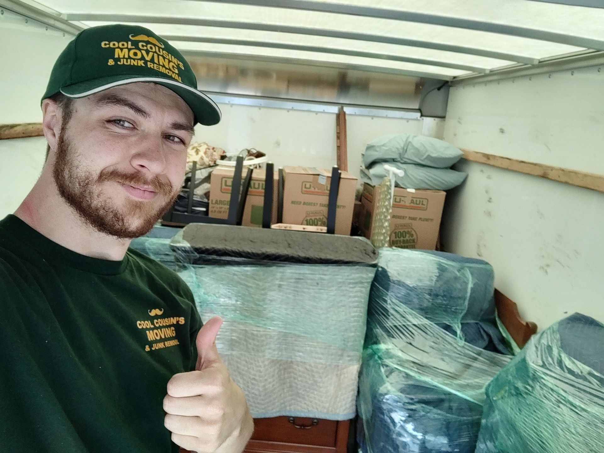 Man in green cap and shirt giving a thumbs-up beside wrapped furniture in a moving truck