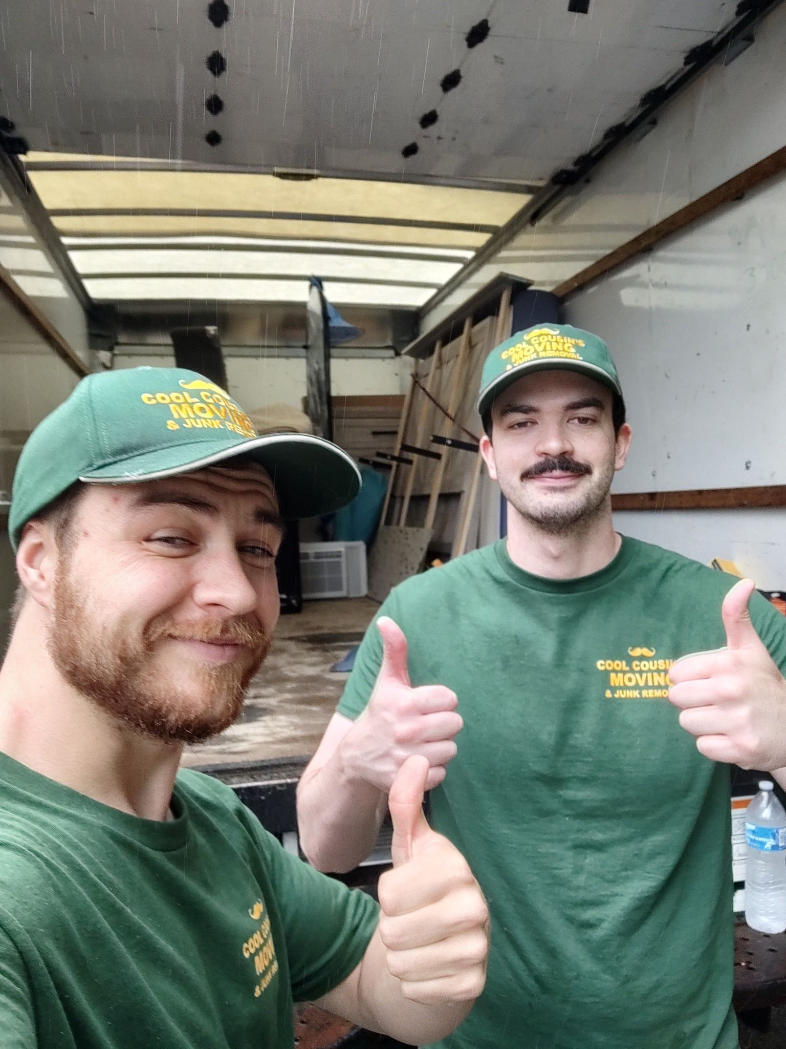 Two men in green shirts and caps giving thumbs up inside a moving truck trailer.