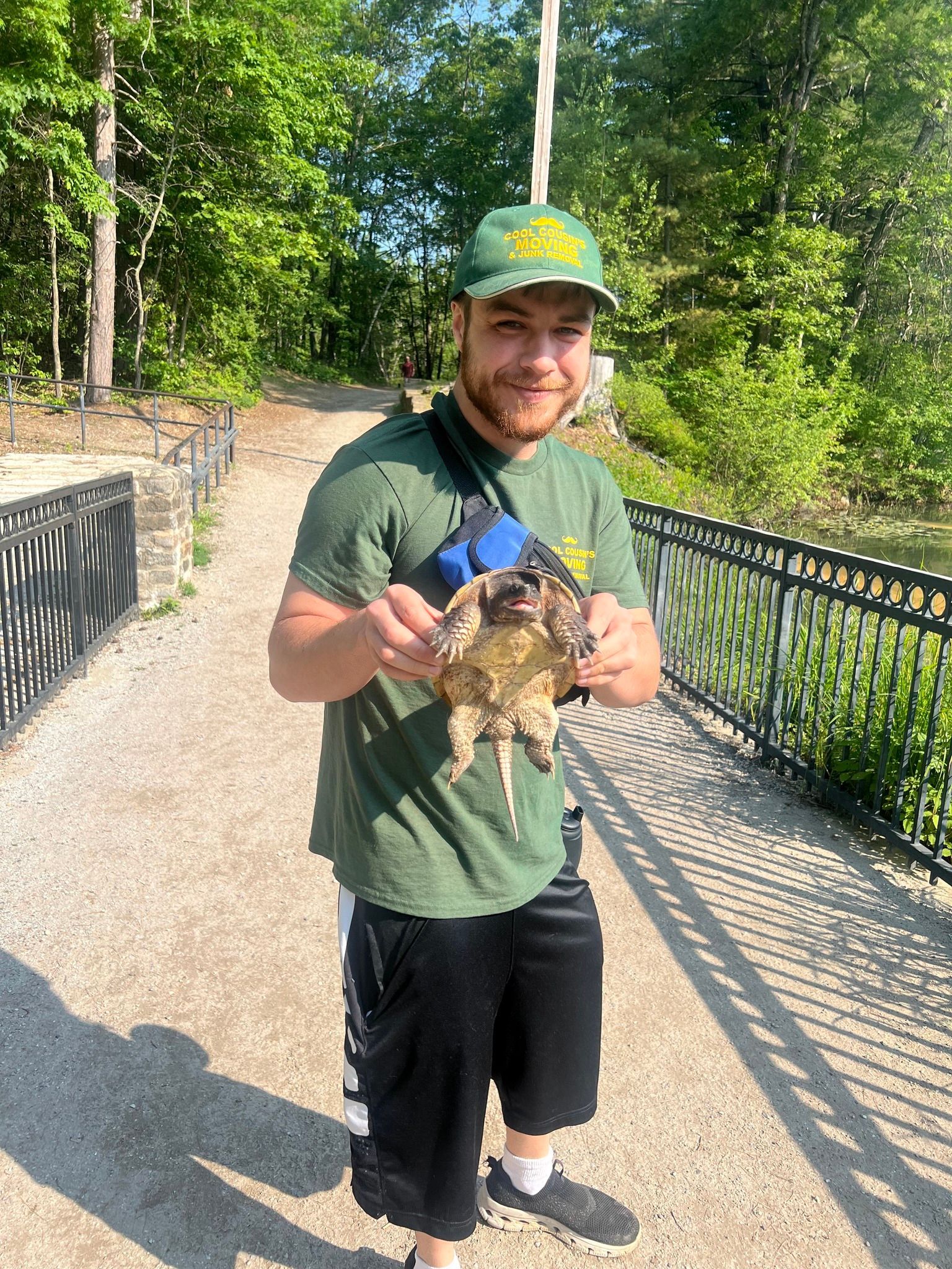 Smiling man holding a cat on a sunlit bridge with trees in the background