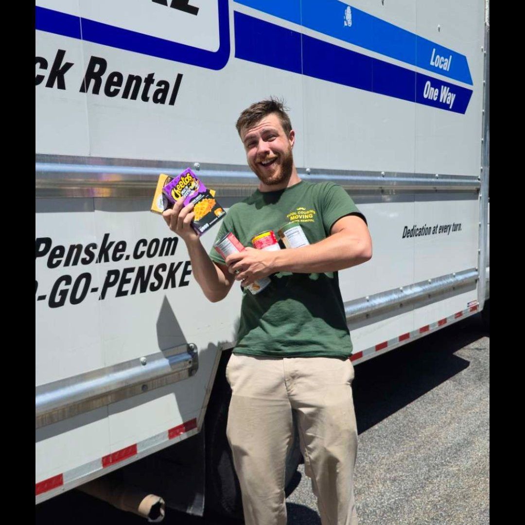 Man smiling beside a Penske moving truck, holding colorful boxed items outdoors.