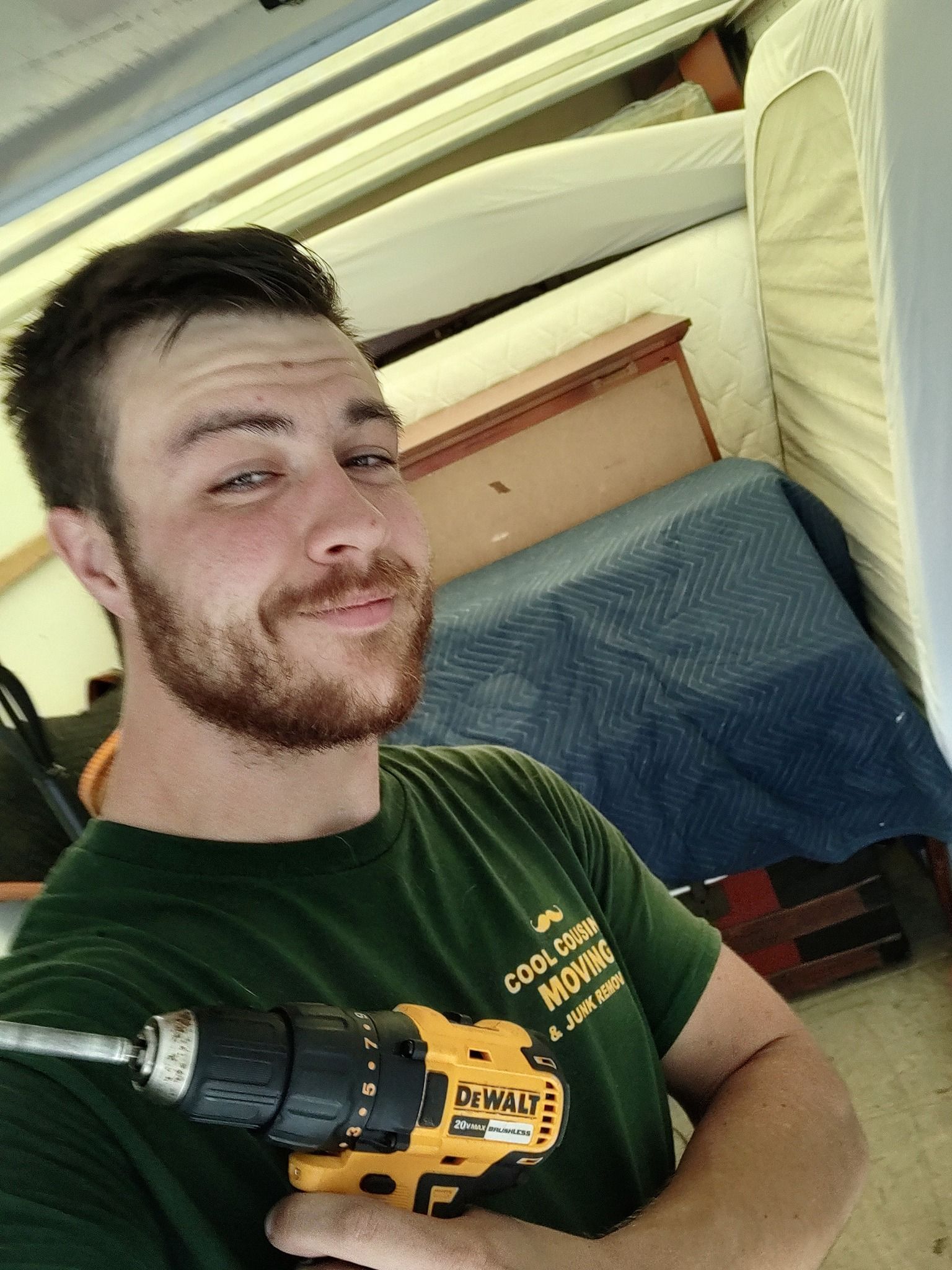 Person holding a yellow power drill while standing in a cluttered room with stacked mattresses.