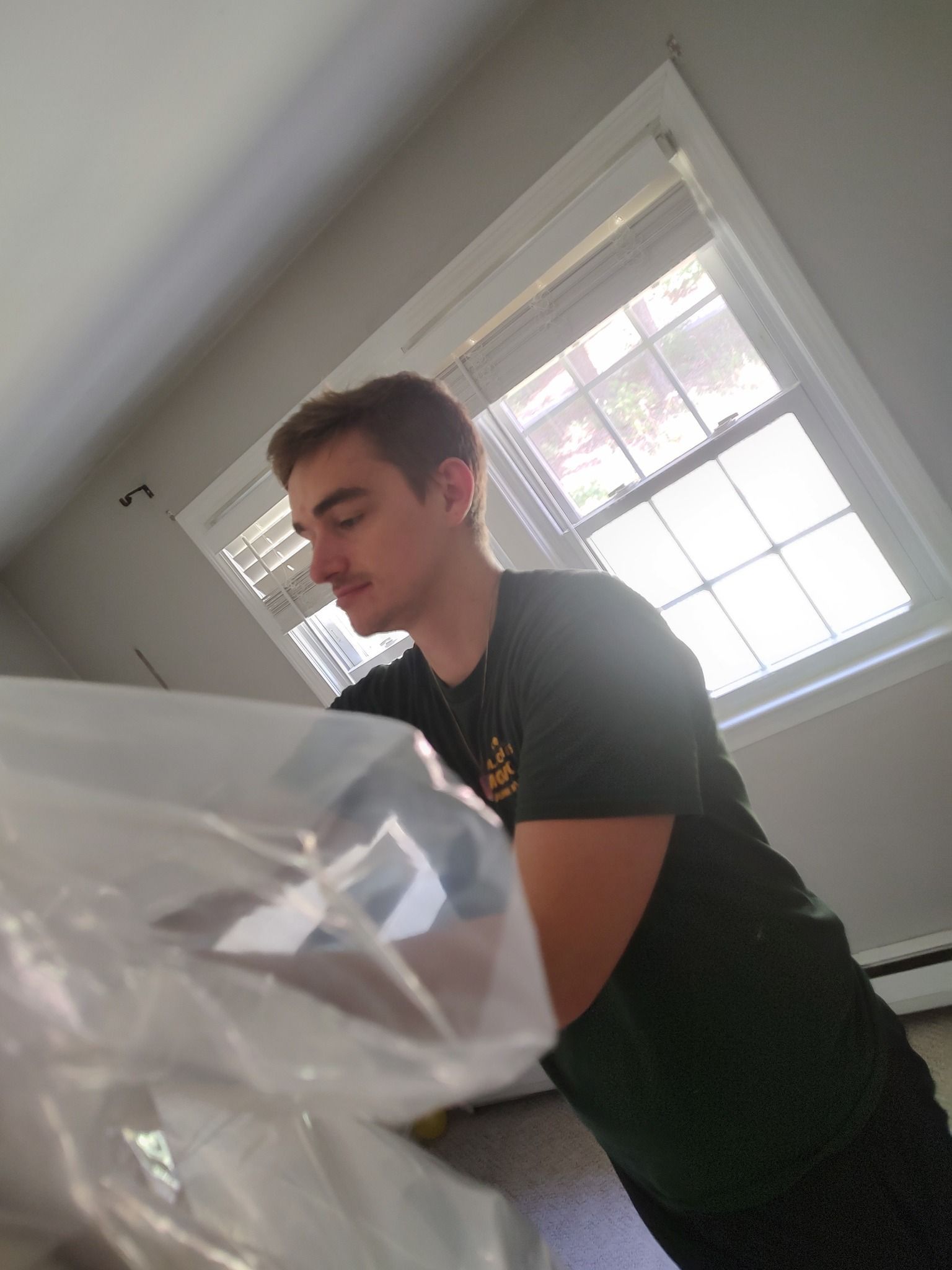 Person in a black shirt leaning over a plastic-covered object near a bright window.
