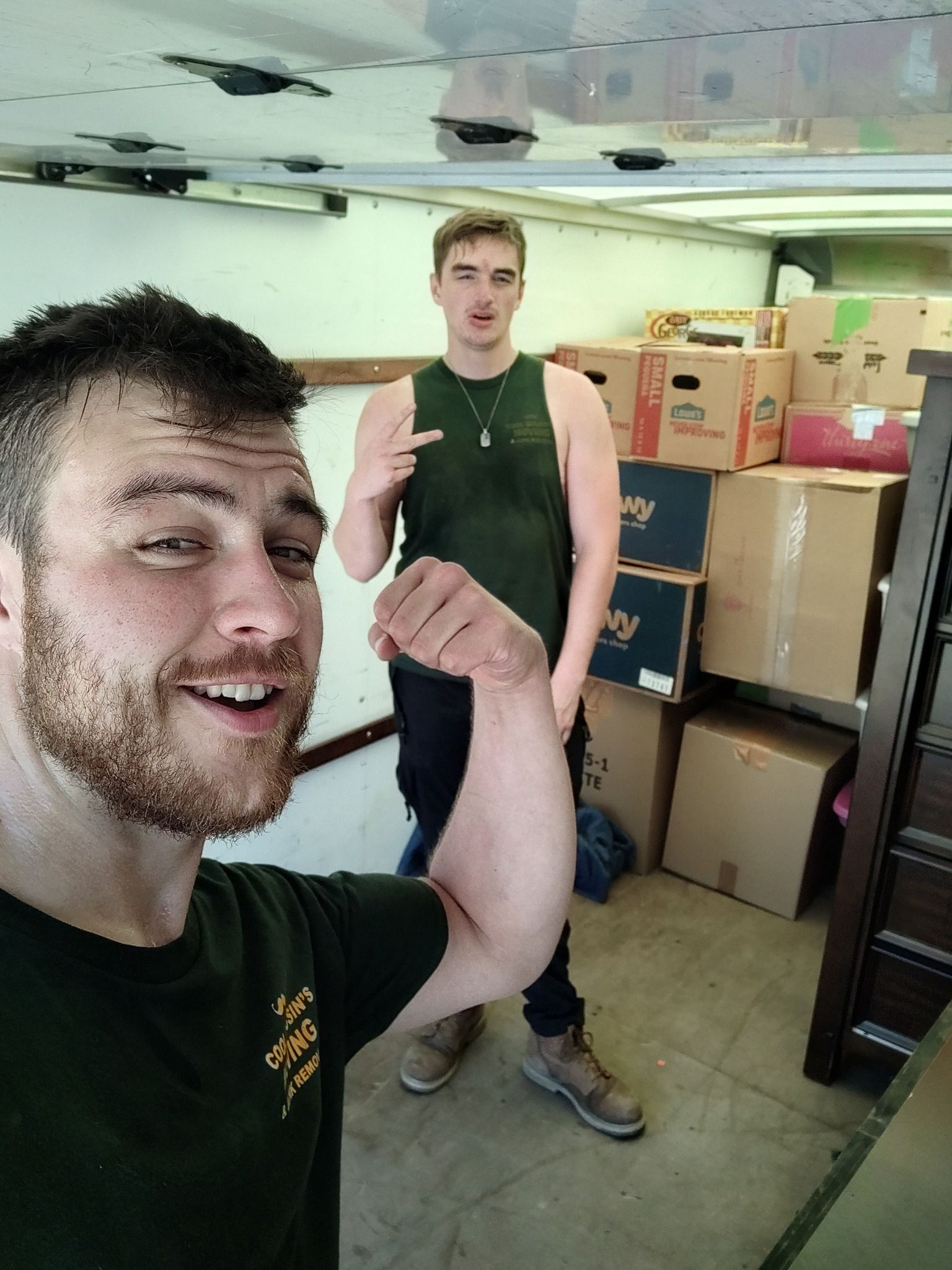 Two men in a moving truck, one taking a selfie with stacked cardboard boxes behind them
