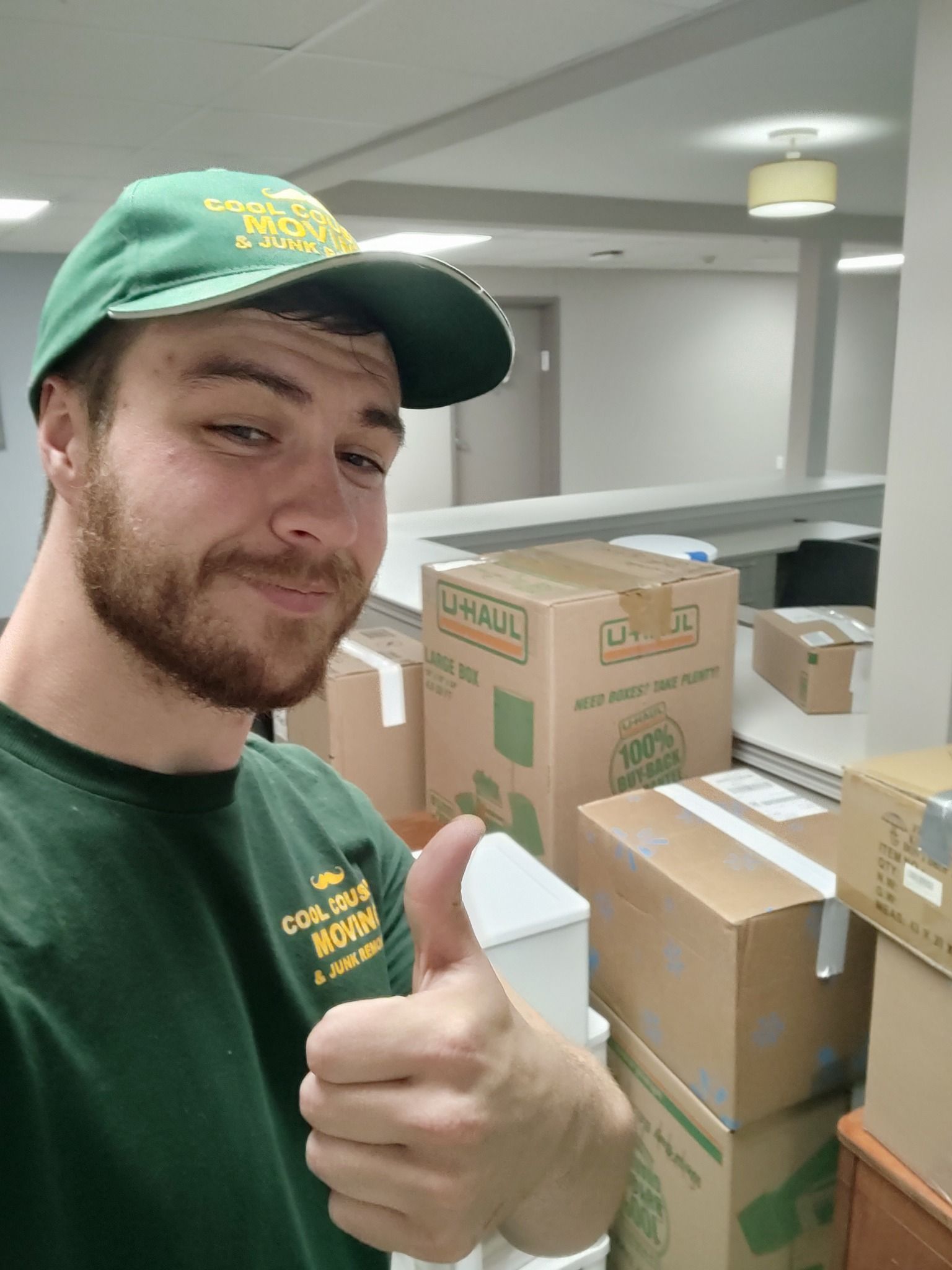 Smiling man in green cap and shirt gives a thumbs-up beside stacked shipping boxes in a warehouse.