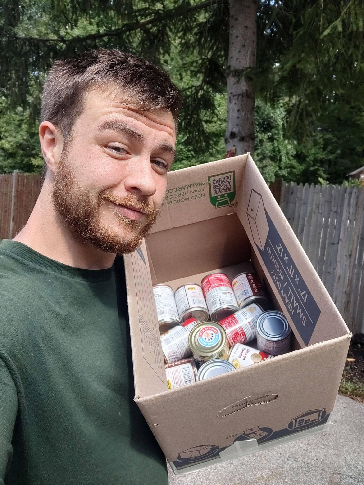 Man holding a box of canned food outdoors, smiling at the camera.