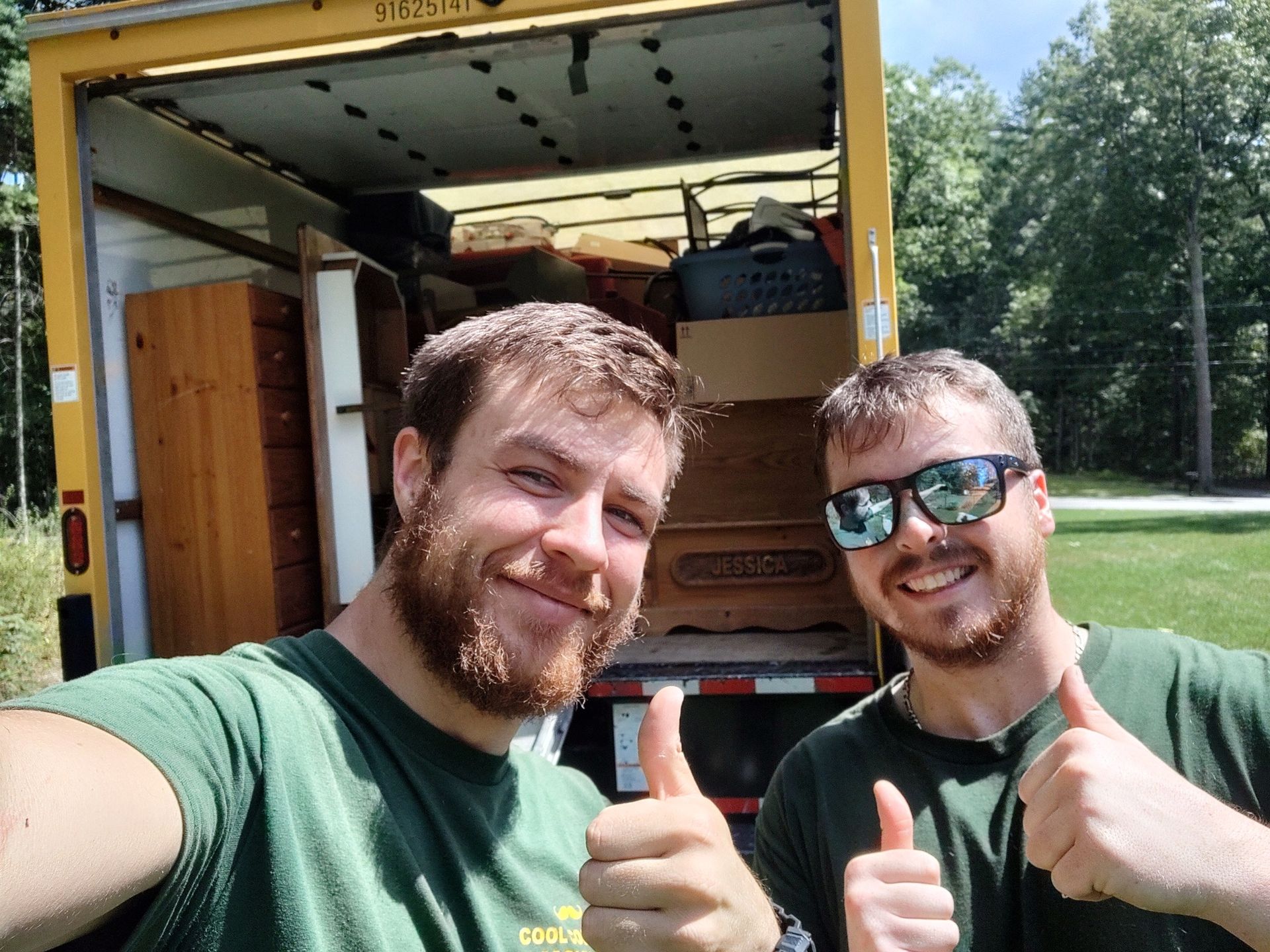 Two smiling men giving thumbs up in front of an open truck in a grassy park