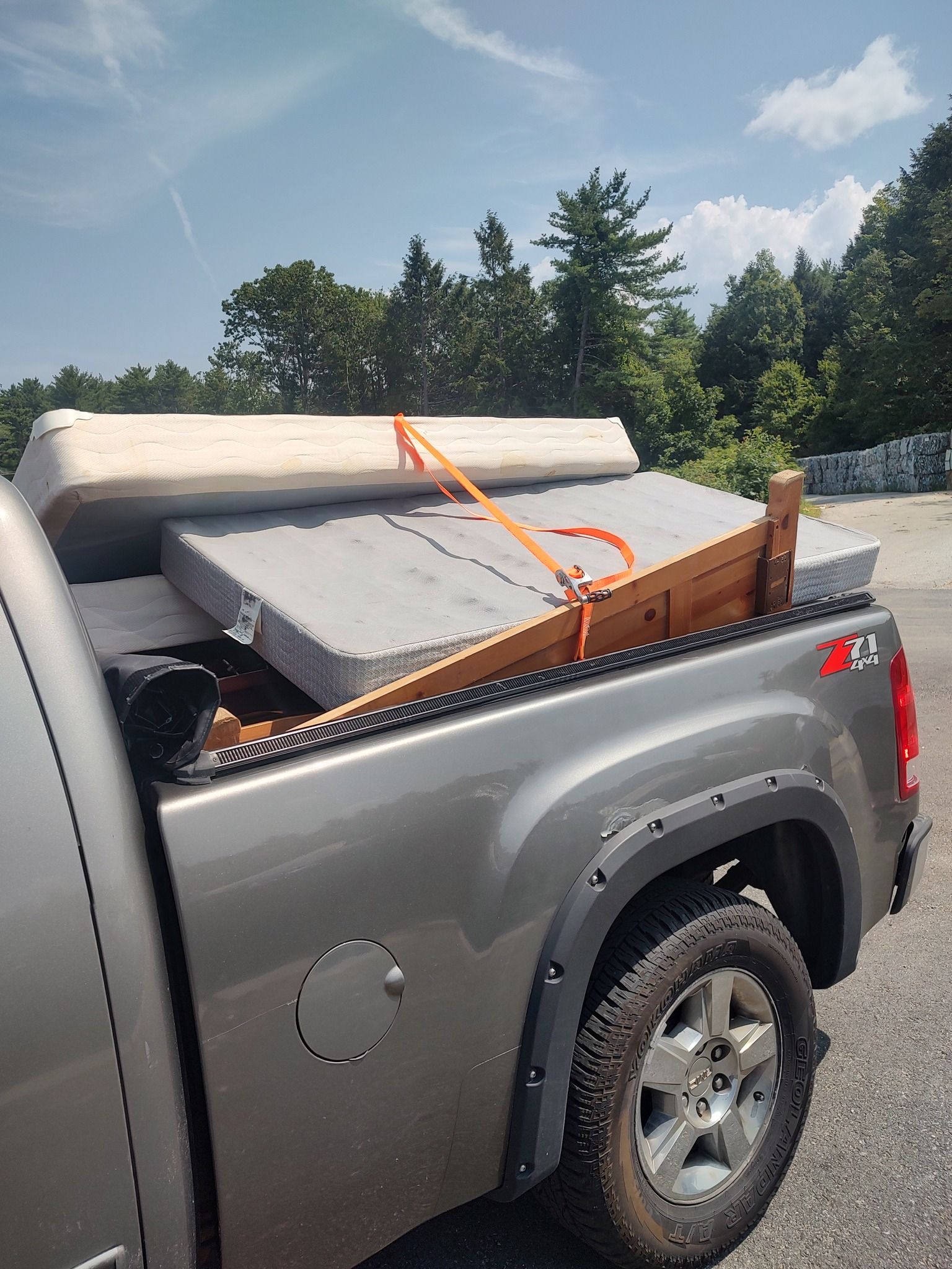 Gray pickup truck loaded with foam cushions and a mattress, secured with orange straps outdoors under a blue sky