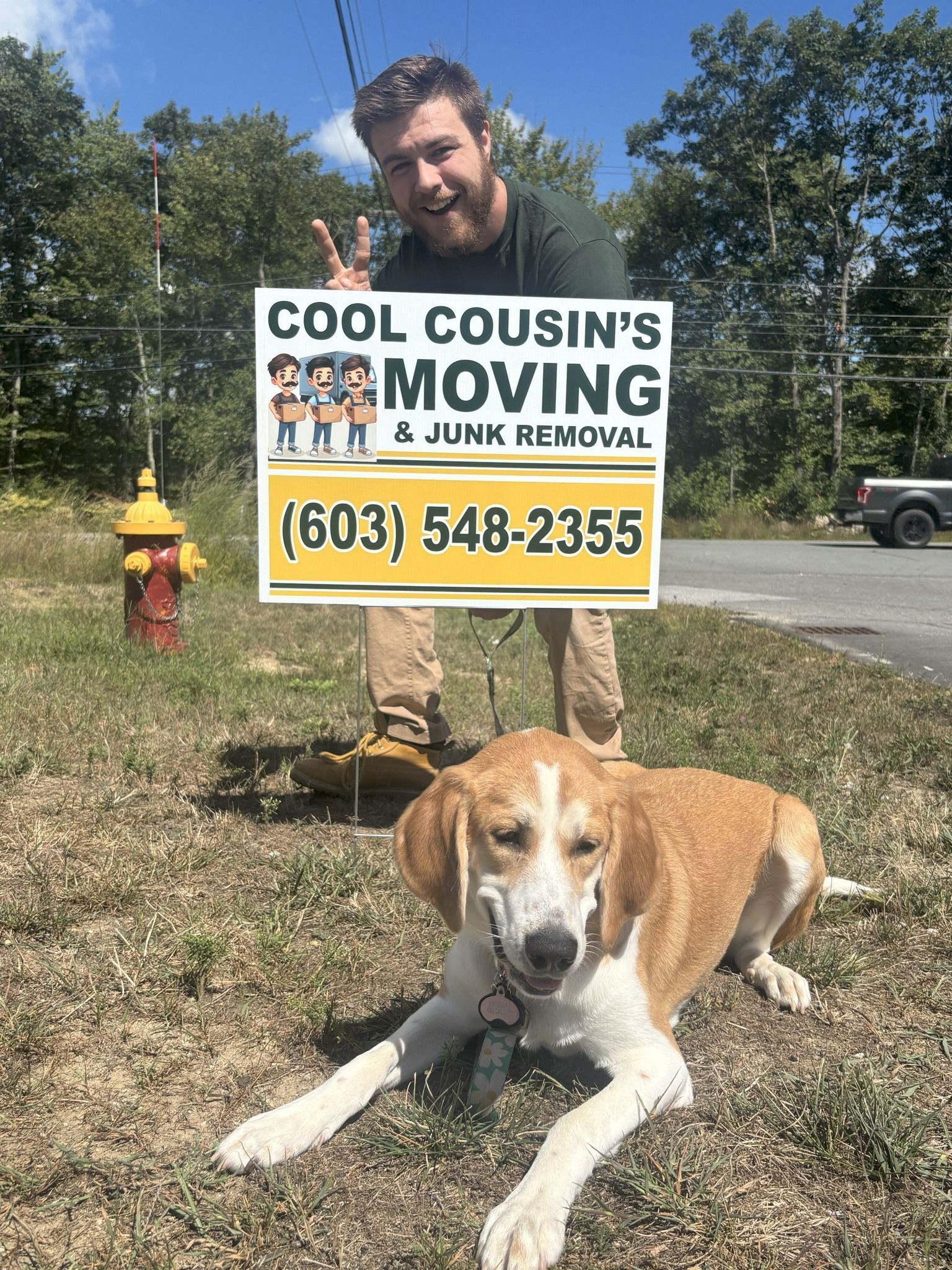 Man holding a “Cool Cousin Moving” sign beside a brown-and-white dog lying on grass outdoors