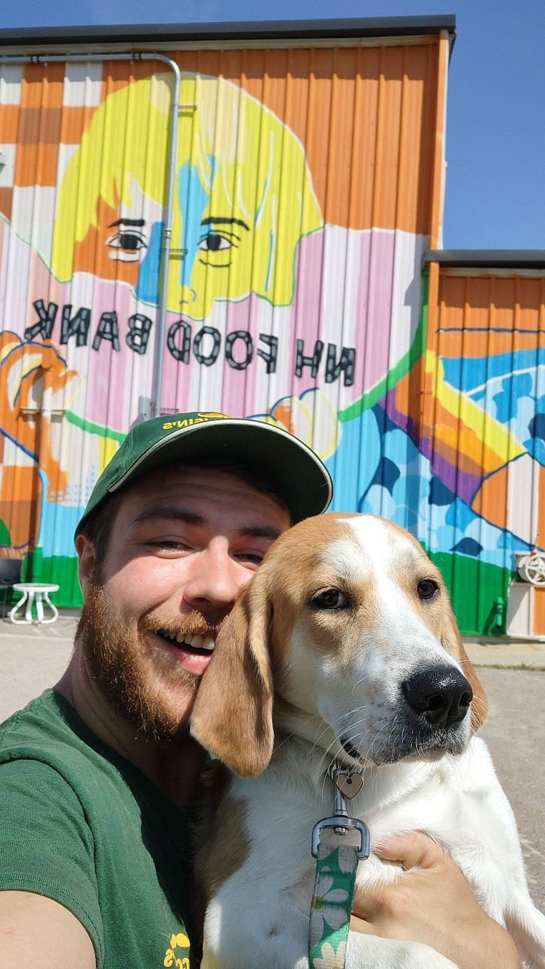 Smiling man in a green cap holds a beagle in front of a colorful mural on a building
