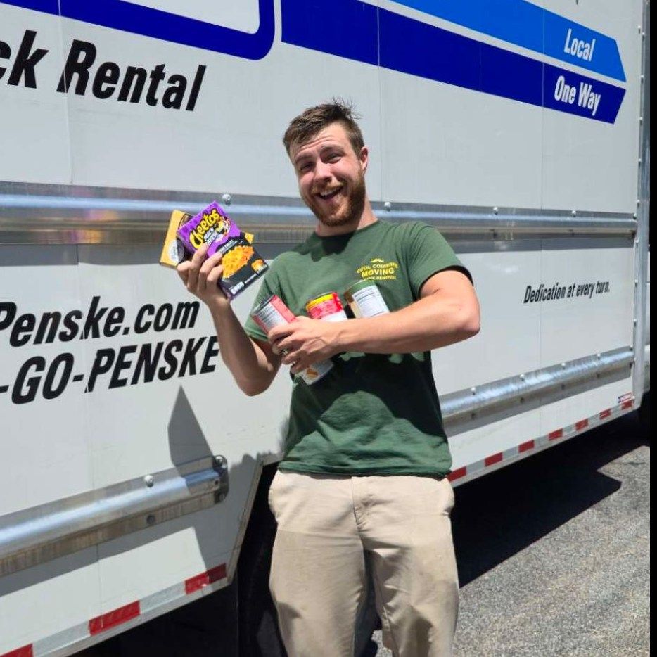 Smiling man in green shirt holding colorful packages beside a Penske rental truck
