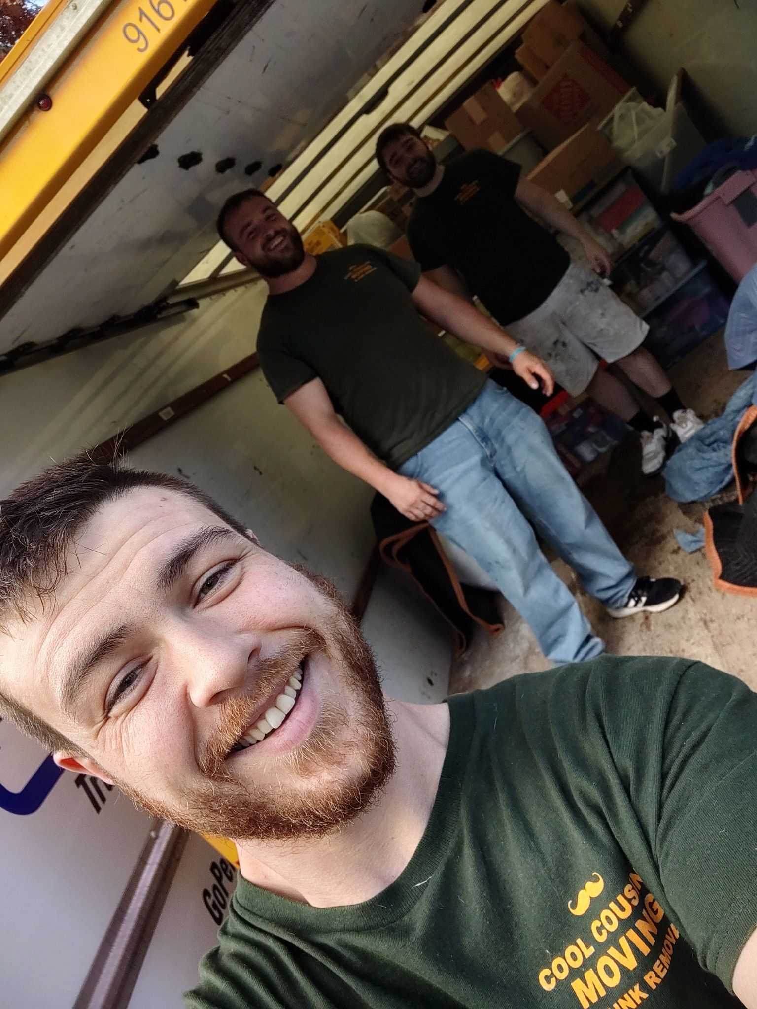 Two smiling men standing in a workshop, one taking a selfie in front of tools and shelves.