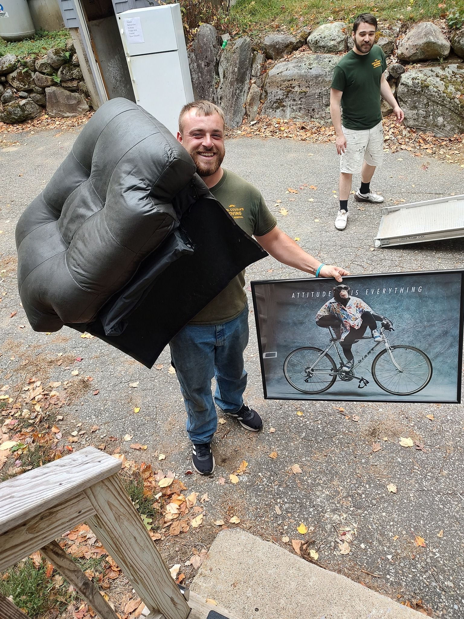 Man holding a black couch and a damaged glass panel in a rocky outdoor area, another person standing behind him