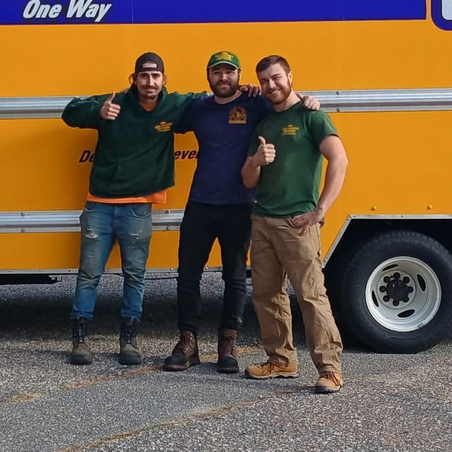 Three men pose and give thumbs up in front of a yellow moving truck.
