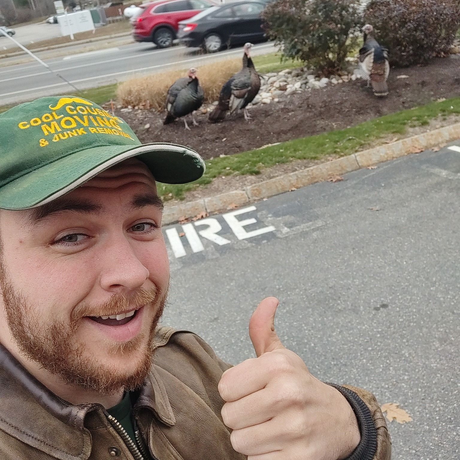 Smiling man in green cap gives thumbs-up near wild turkeys in a parking lot.