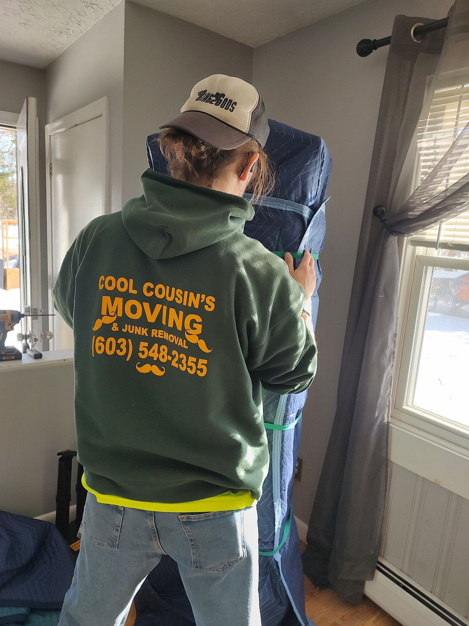 Person in a green hoodie and cap taking a mirror selfie in a bright room.