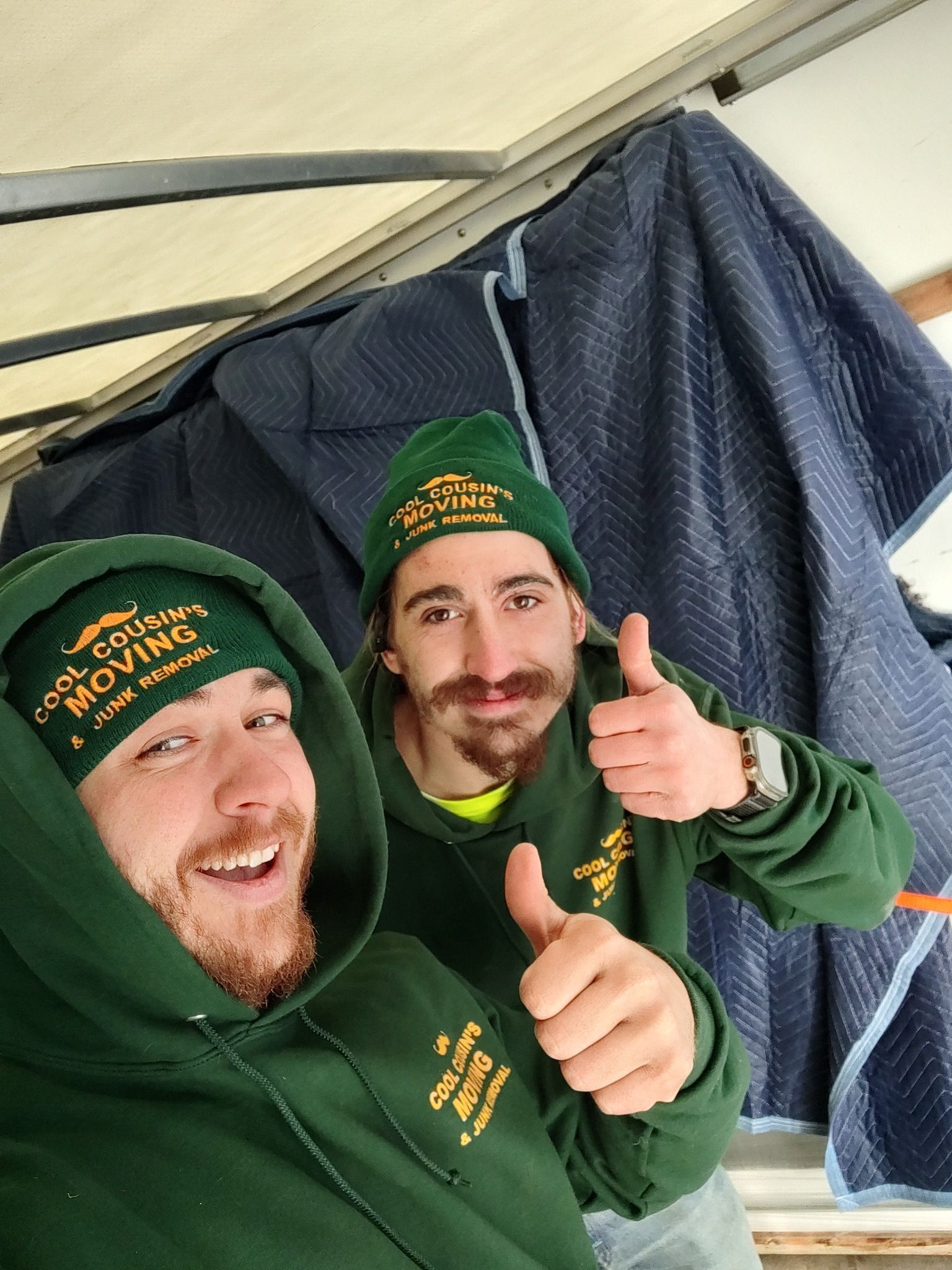 Two men in green hoodies and hats smiling and giving thumbs up in front of a dark tarp backdrop