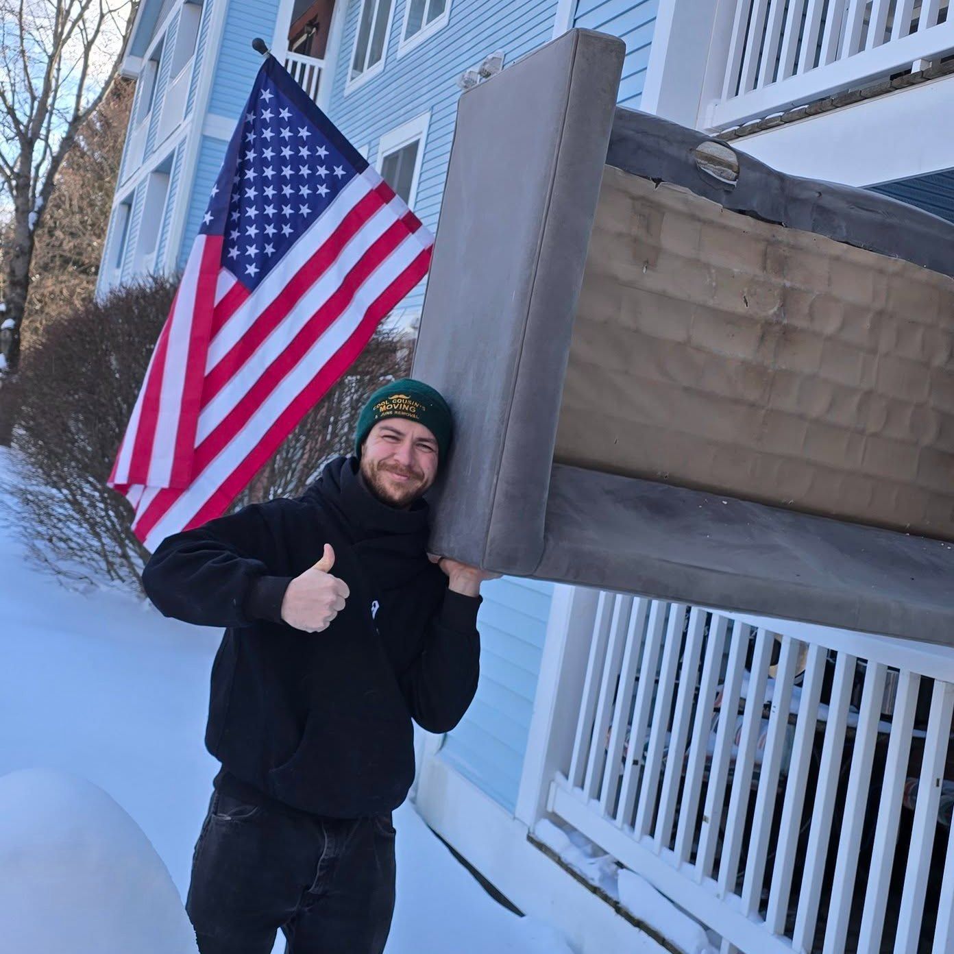 Person smiling and giving a thumbs-up beside a large American flag in a snowy outdoor setting.