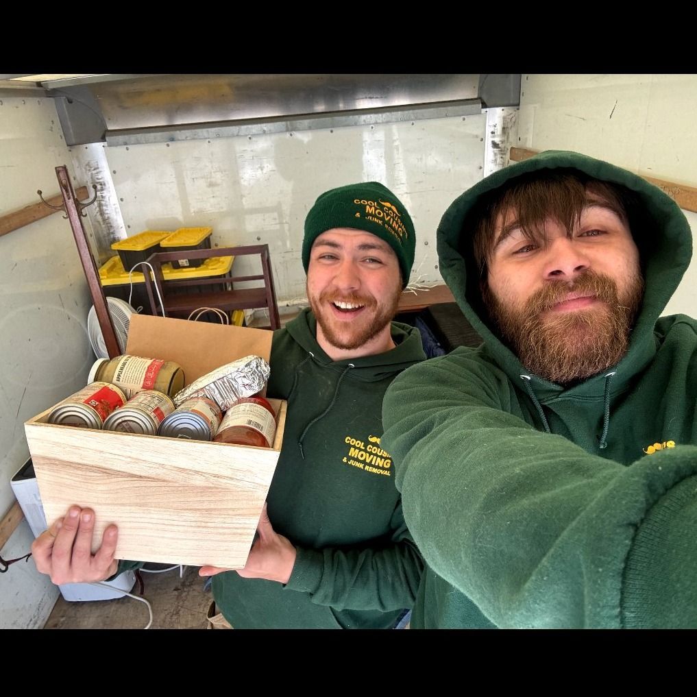Two smiling workers in green hoodies holding a wooden box of canned goods inside a van