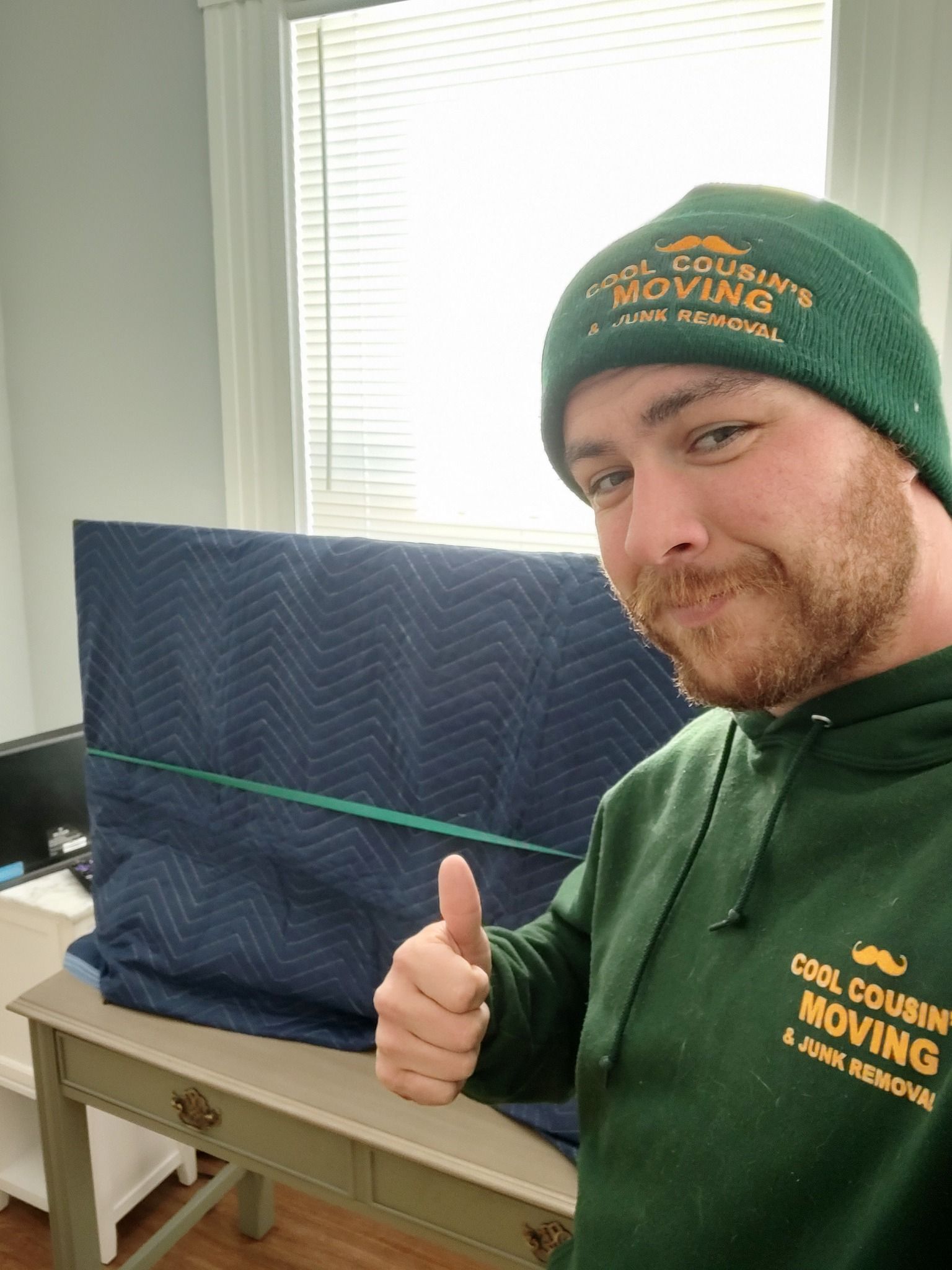 Man in green beanie and hoodie giving thumbs up beside a wrapped item in a bright room.