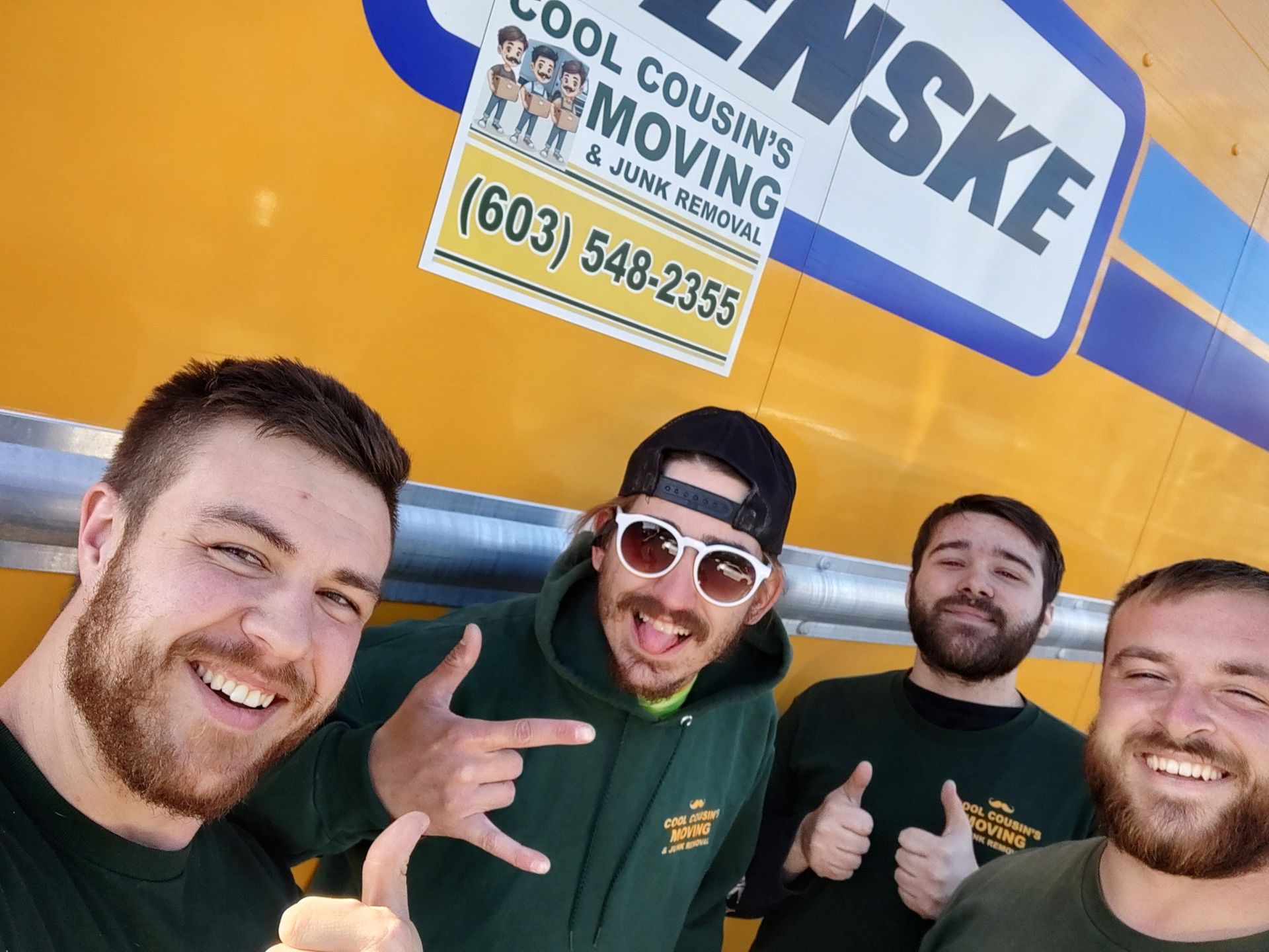 Four smiling men posing in front of a yellow Penske moving truck sign, giving thumbs-up and rock hand gestures