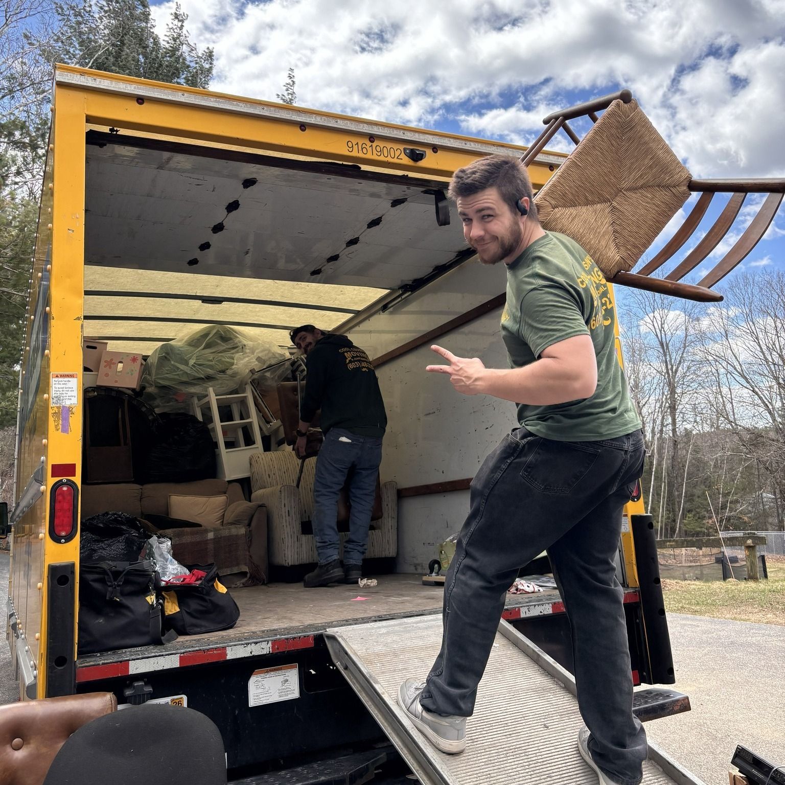 Two men unloading items from a yellow moving truck, one carrying a chair up the ramp
