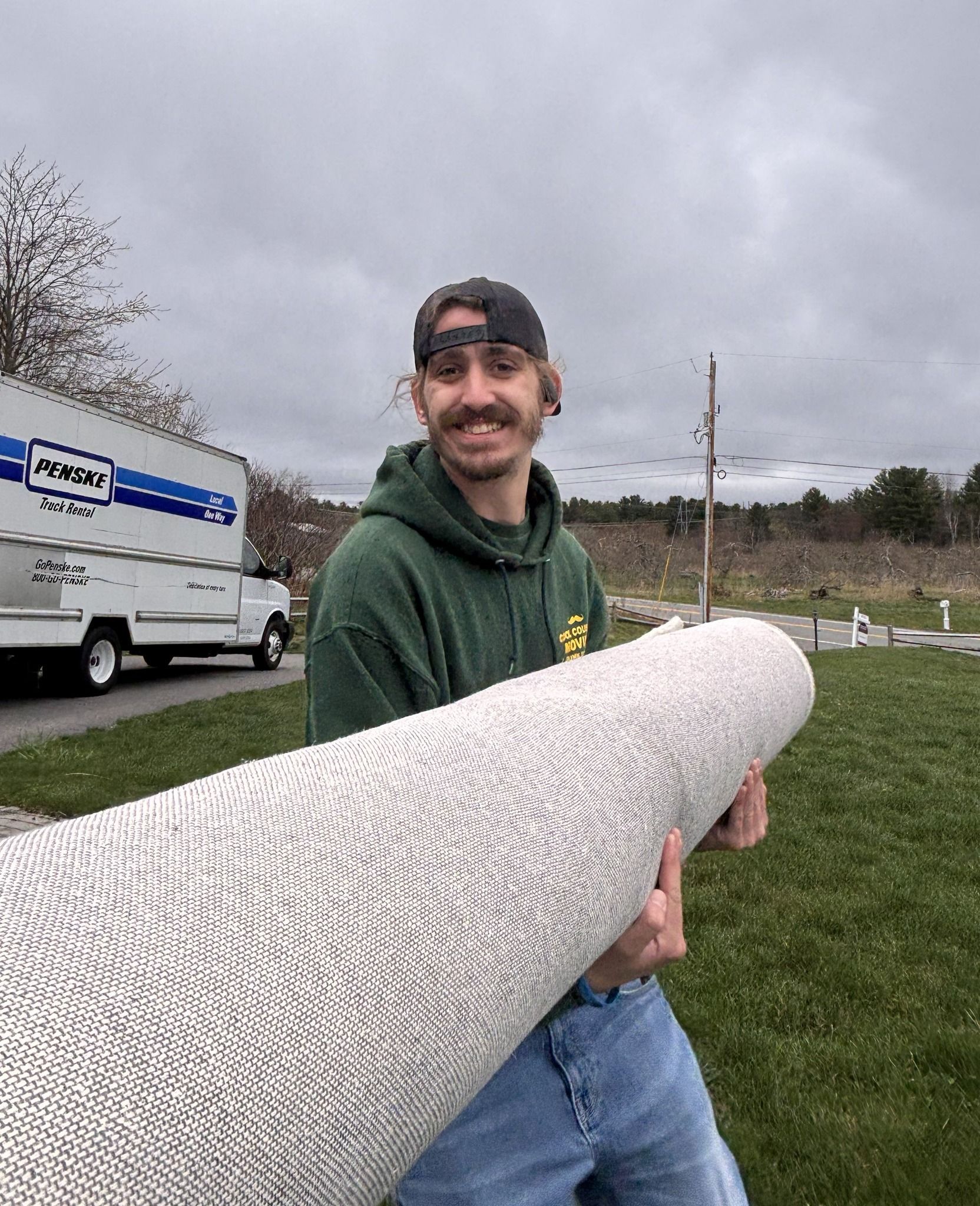 Man holding a rolled white rug outdoors near a moving truck under a cloudy sky.