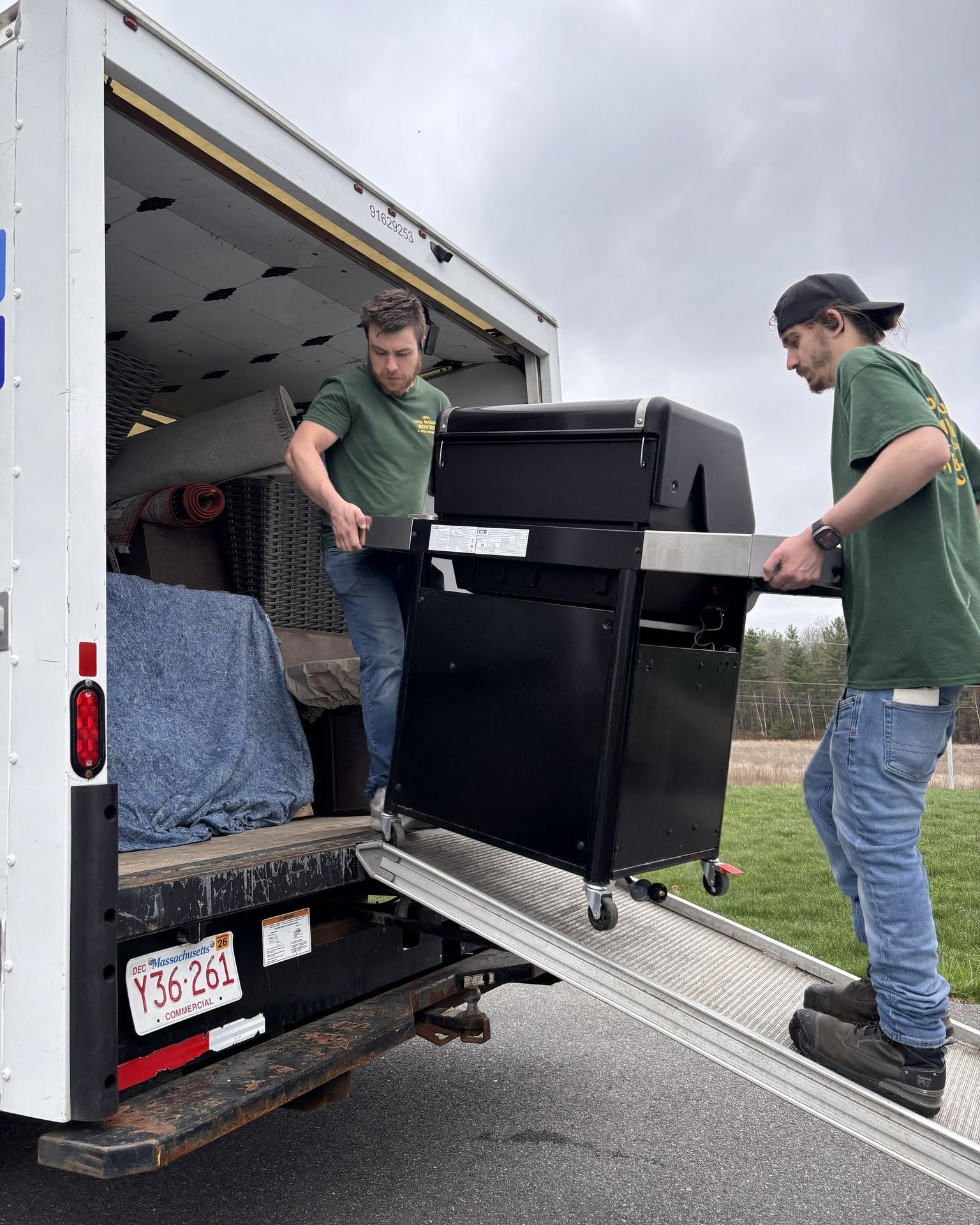 Two movers in green shirts carrying a black cabinet up a ramp into a moving truck.