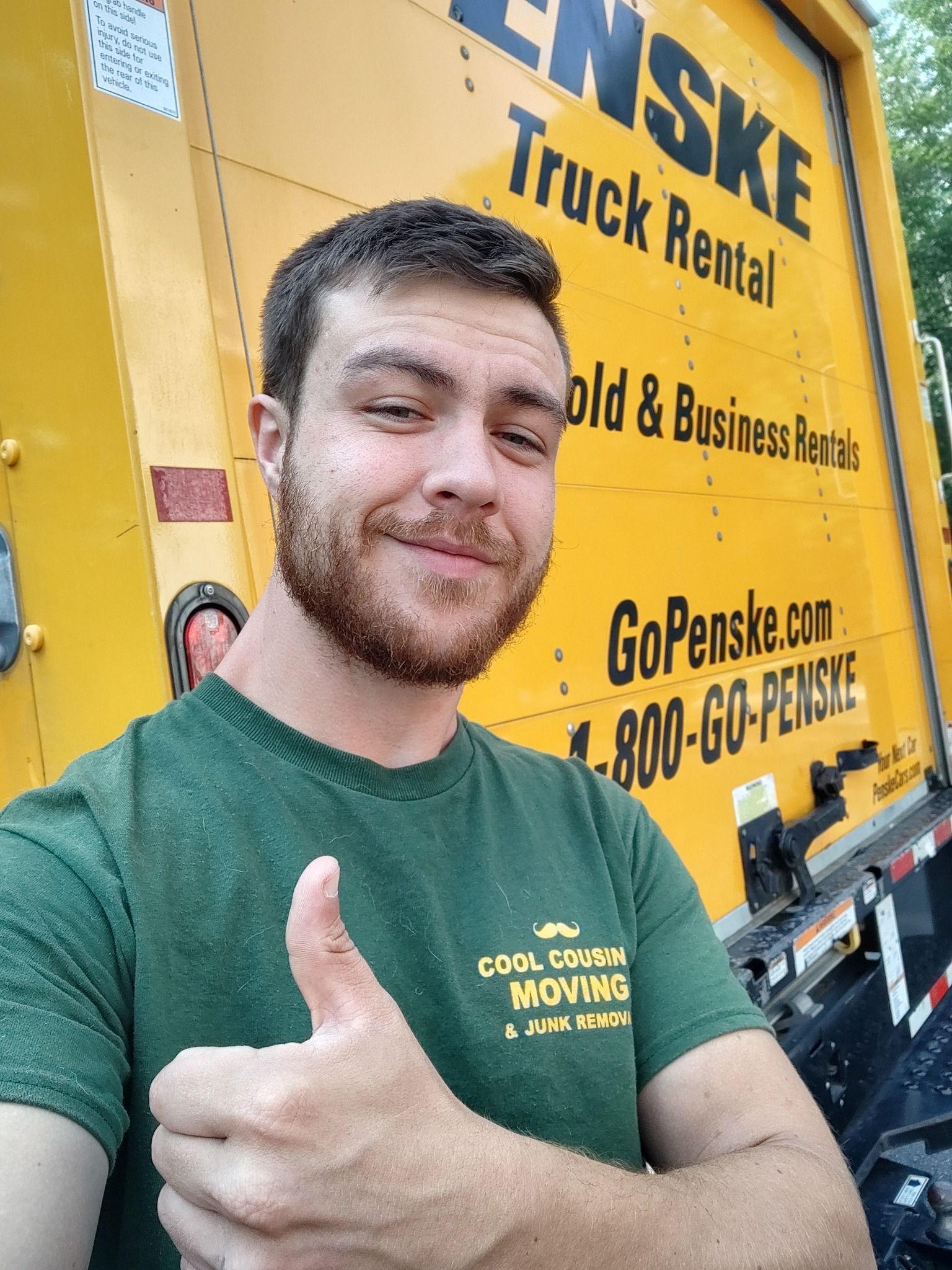 Smiling man in green shirt giving thumbs-up in front of yellow moving truck sign