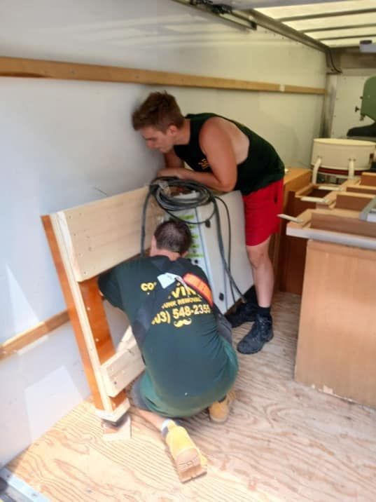 Two people assembling wooden furniture in a workshop, one kneeling with a drill and one leaning over the piece