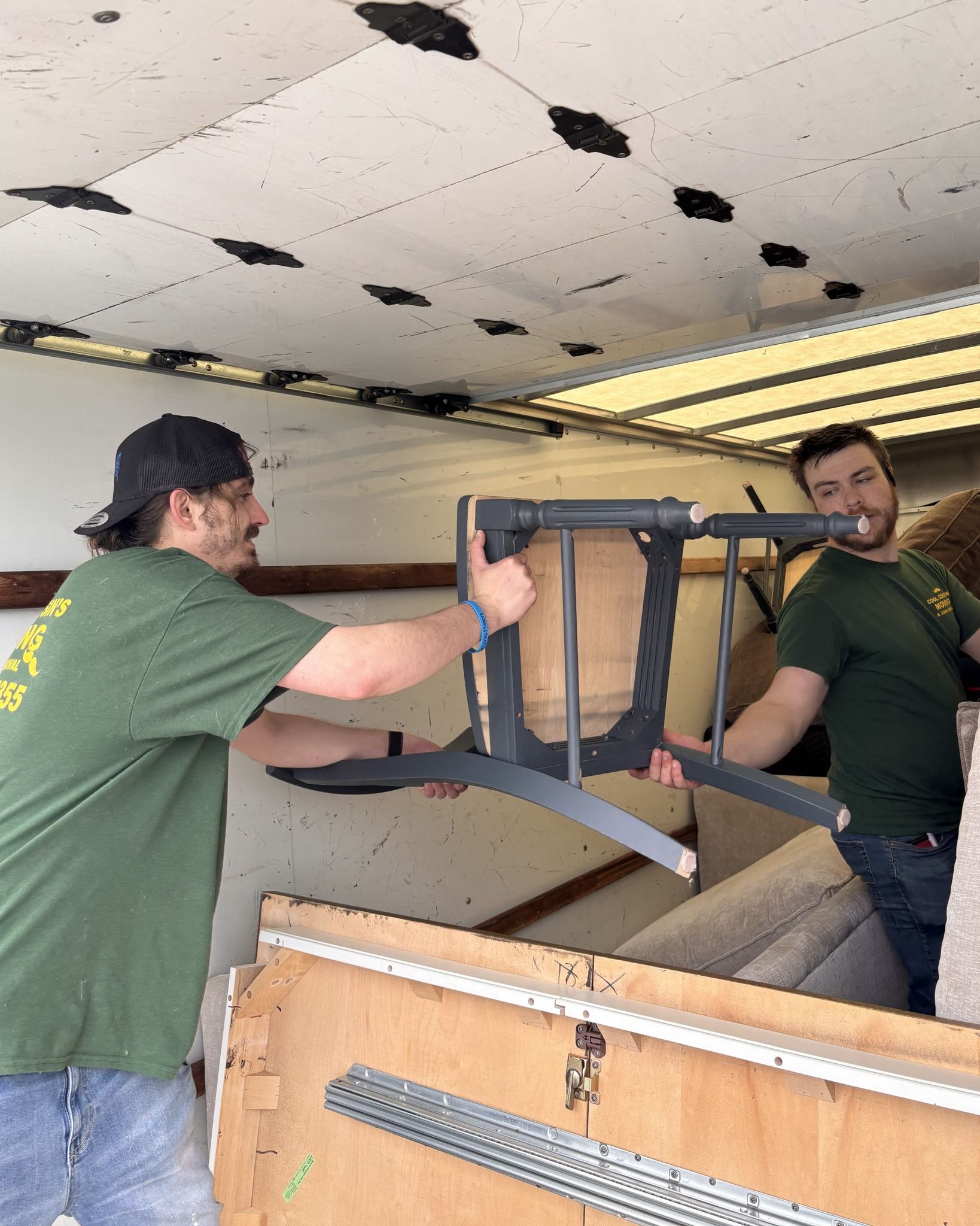 Two people in green shirts carry a chair through a truck or trailer interior.