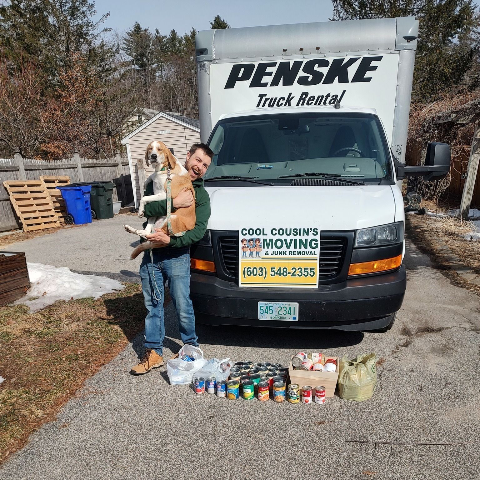 Man holding a dog beside a Penske rental truck with moving supplies and canned donations lined up in front