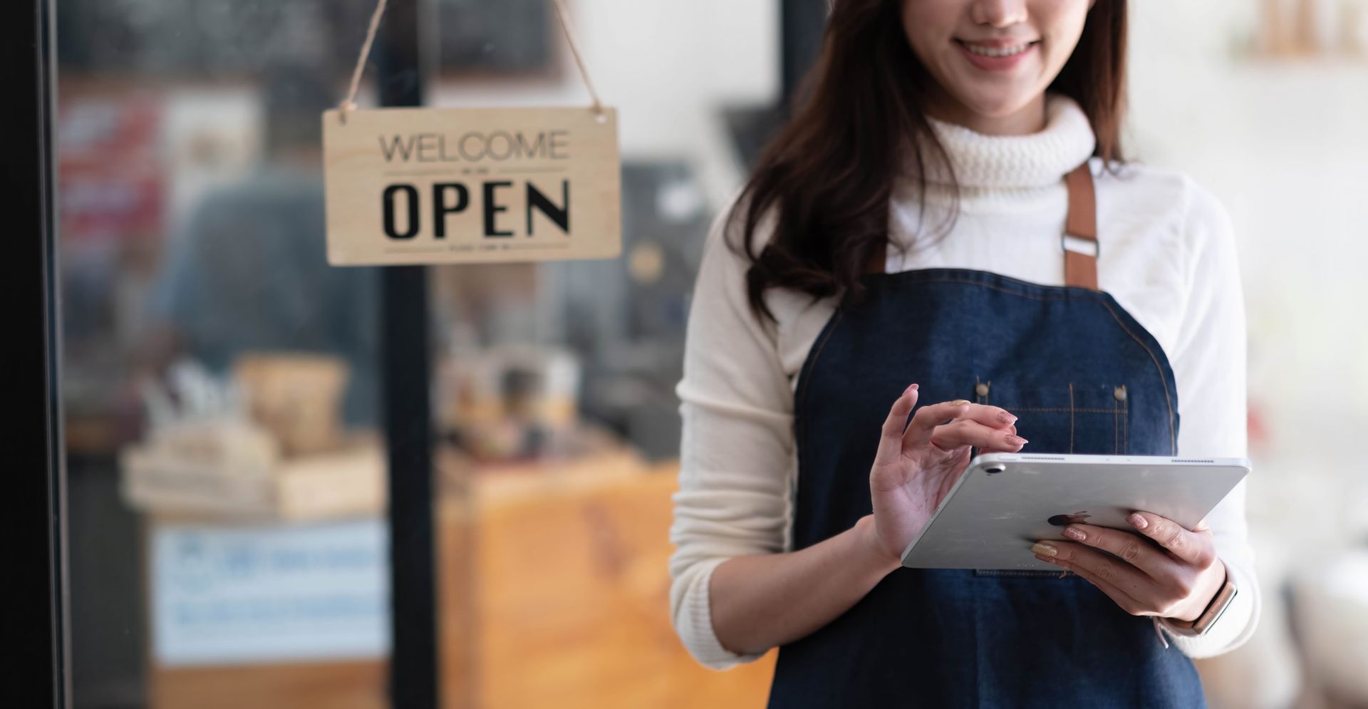 Shopkeeper in apron using a tablet beside a storefront “Welcome Open” sign