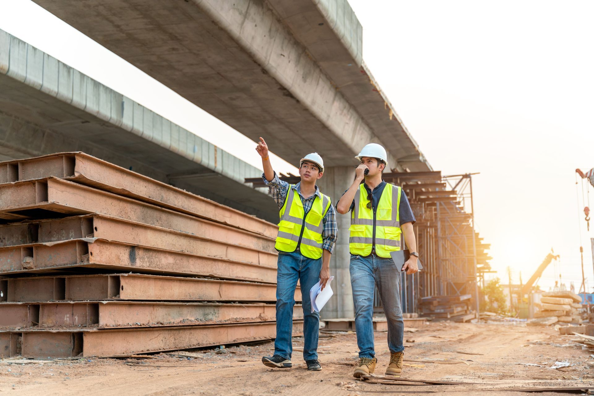 Two construction workers in hard hats and safety vests inspect a bridge site at sunset.