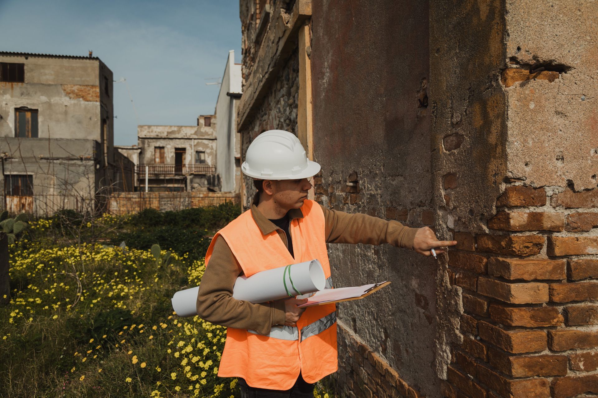 Surveyor in hard hat and orange vest inspecting a brick wall at an old building site.