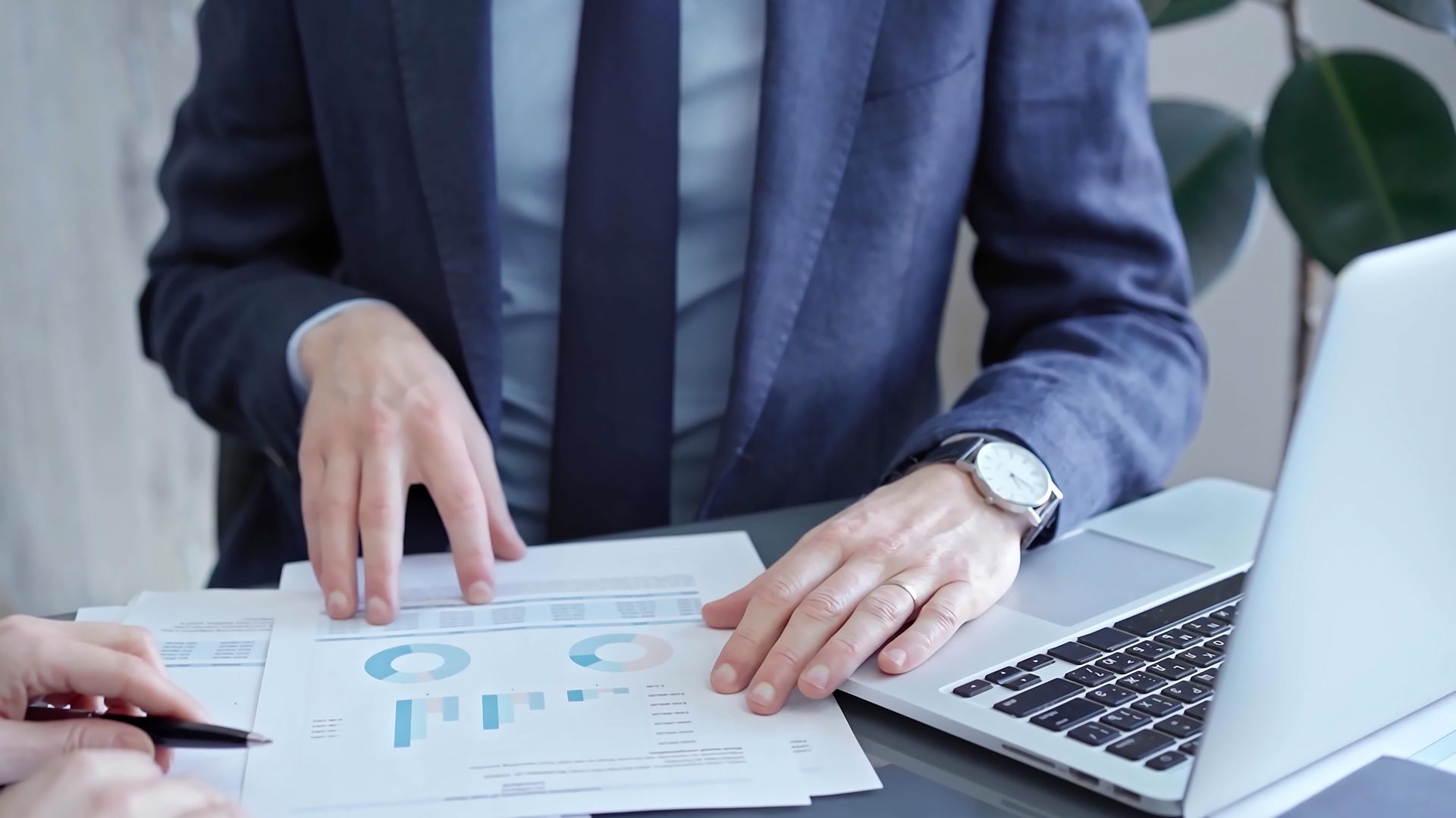 Person in suit pointing at financial graphs with laptop and pen.