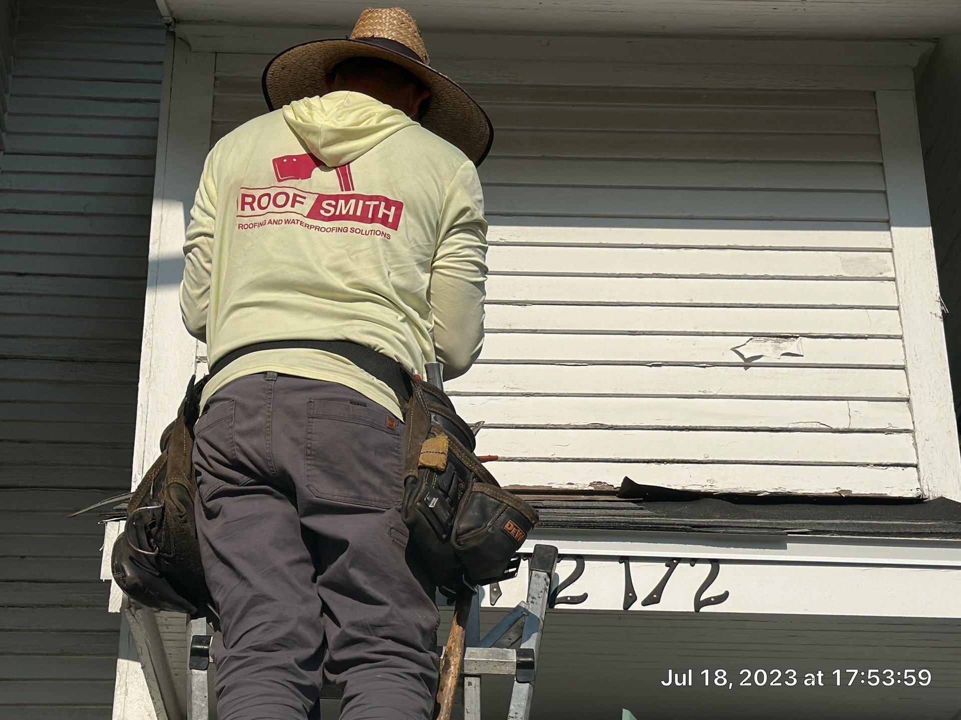 Worker on a roof repairing white siding above a porch, wearing a straw hat and harness.