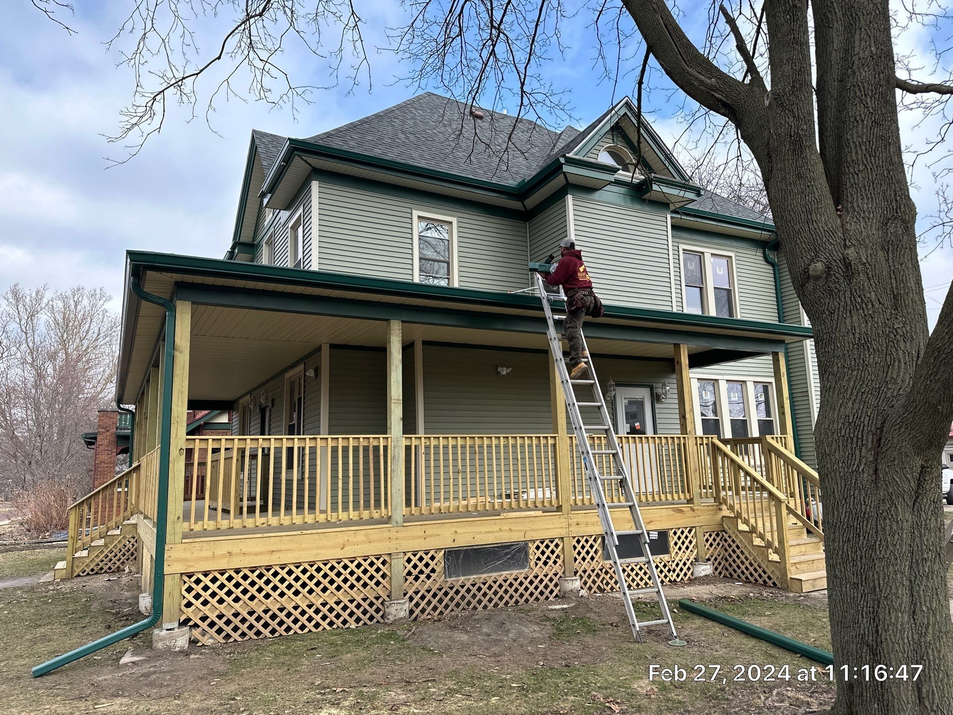 Person on ladder painting trim on a two-story house with green siding and a wraparound porch