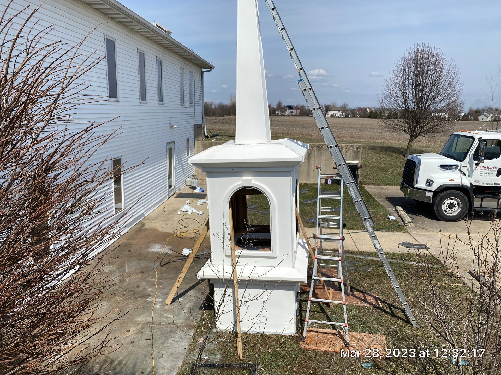 White outdoor fireplace/chimney being installed beside a house, with ladders and a work truck in the yard.