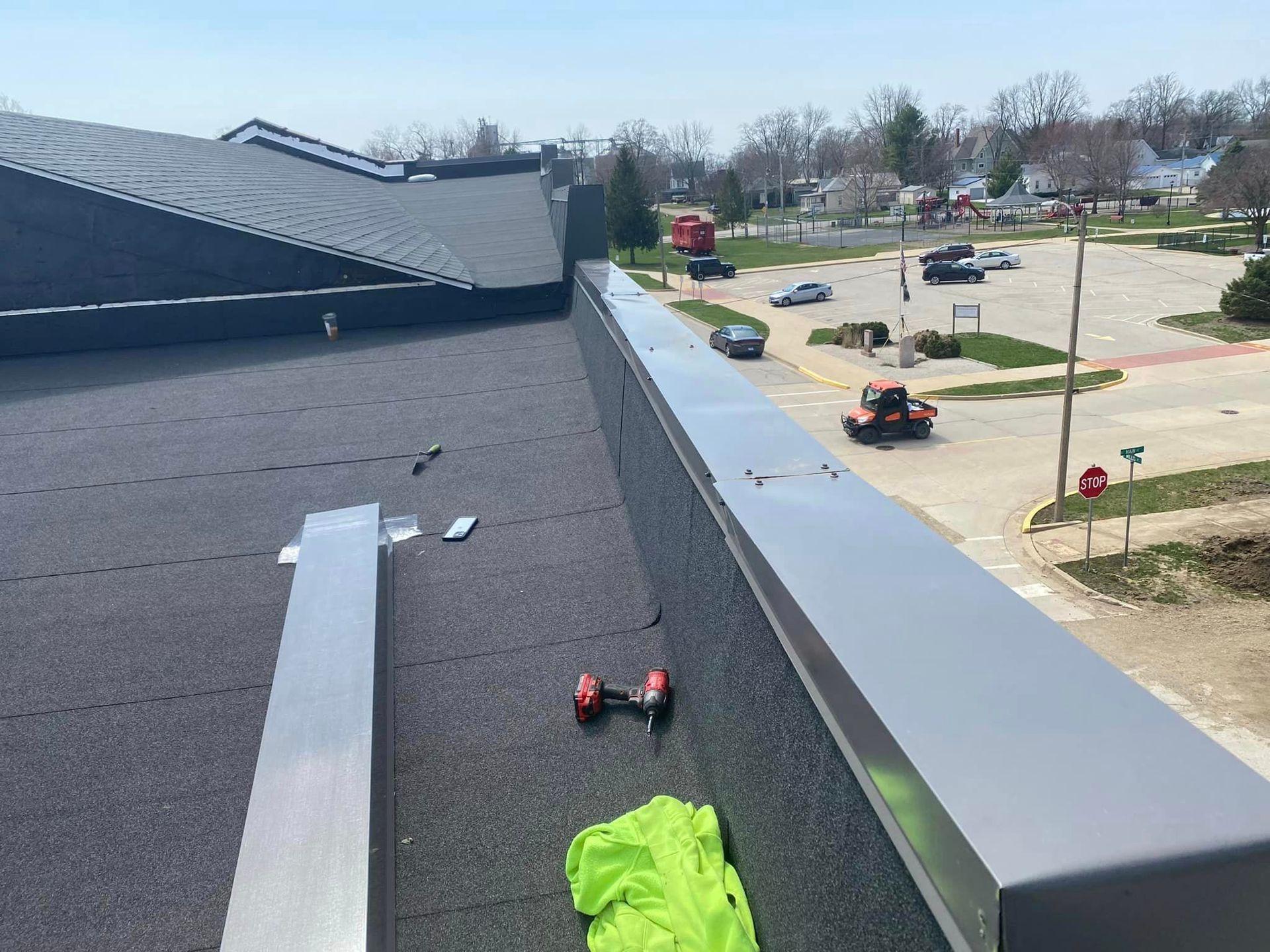 Roof with black shingles and tools near the edge, overlooking a parking lot below.