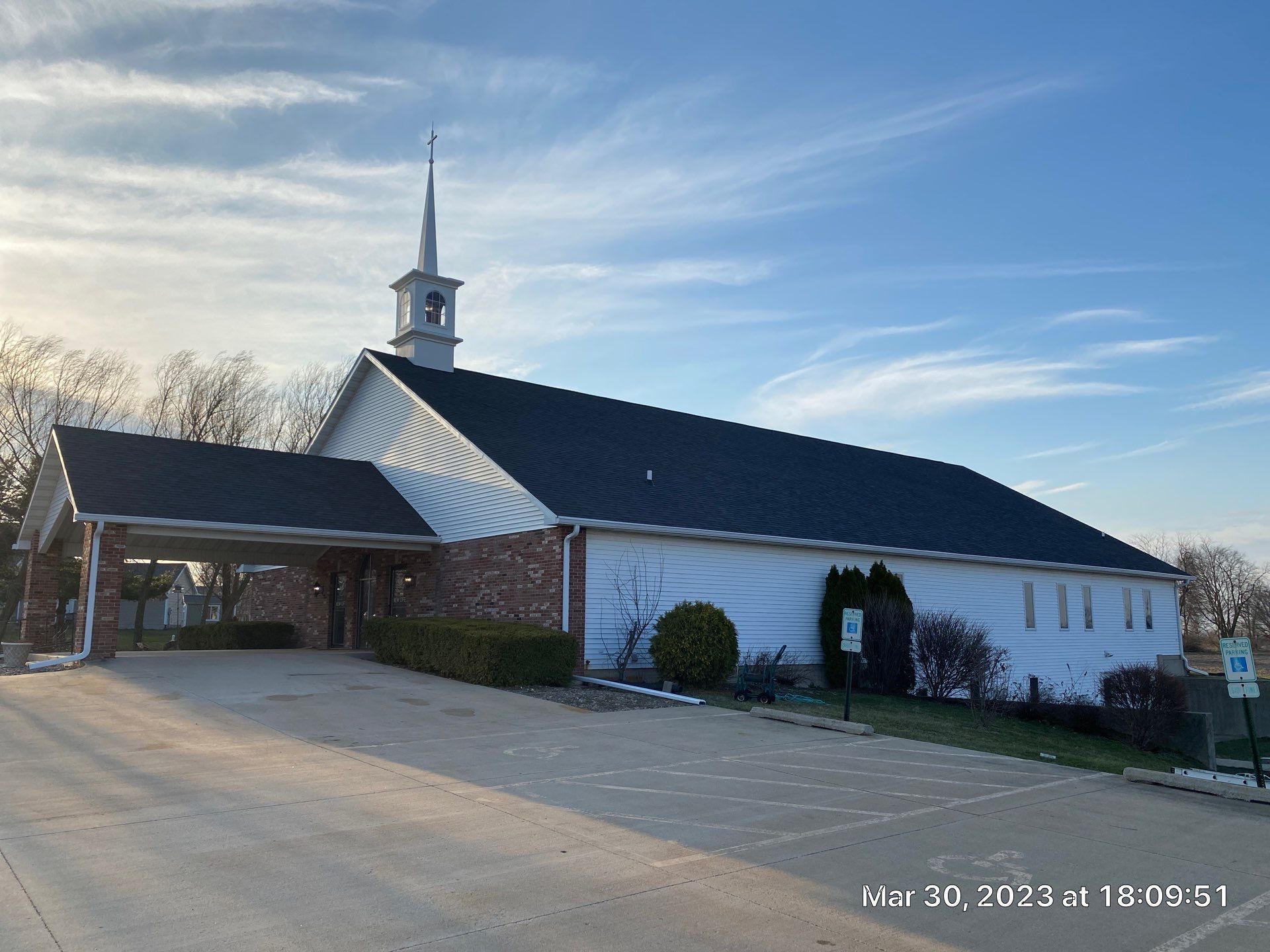 White church with steeple and covered entrance on a sunny day, viewed from the parking lot
